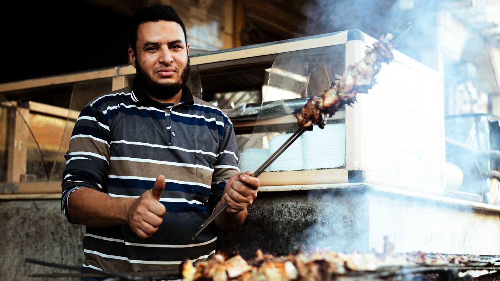 Street vendor holding a grilled meat skewer amid smoke