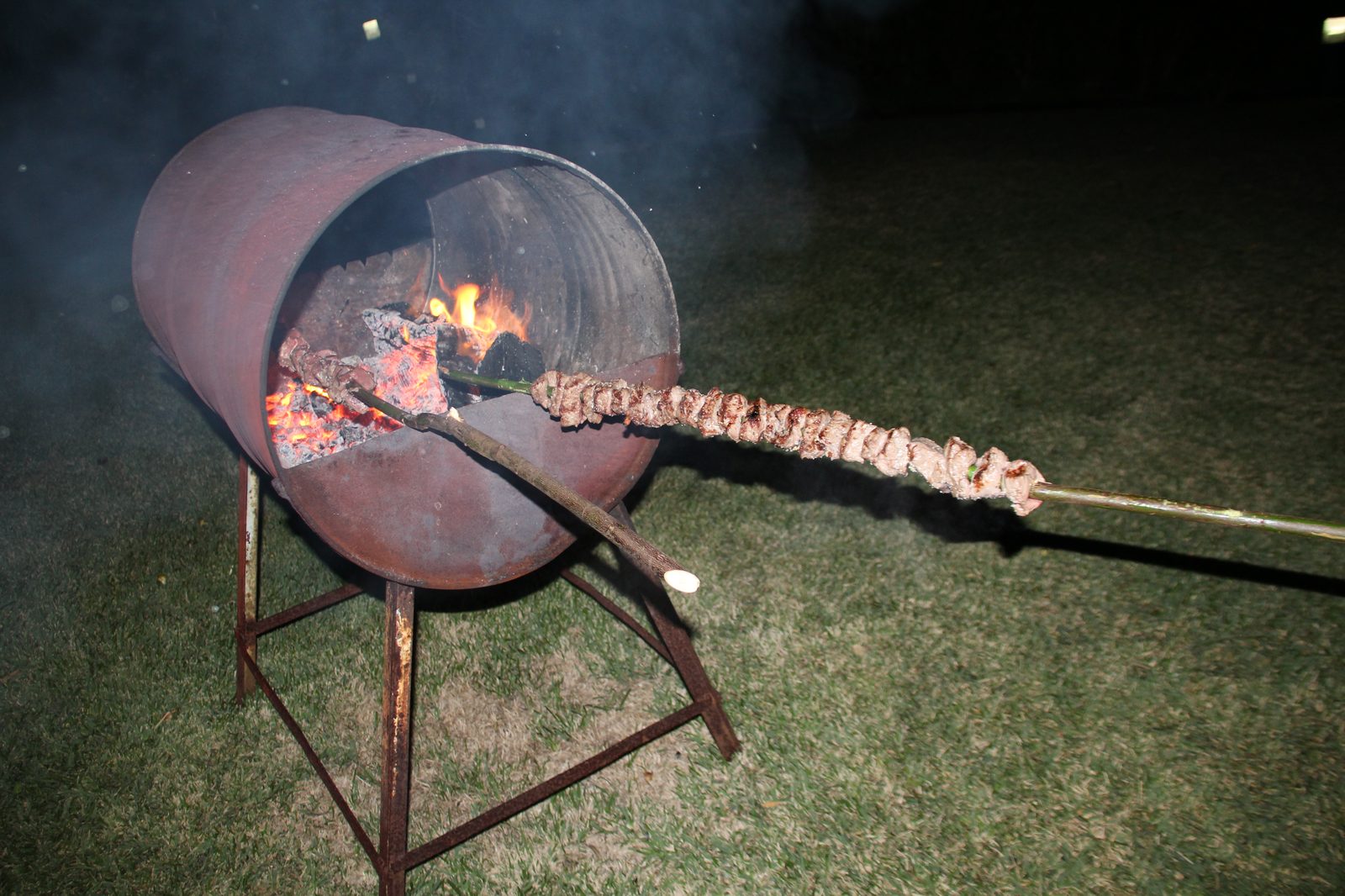 Long meat skewer over a wood-fired barrel grill at night
