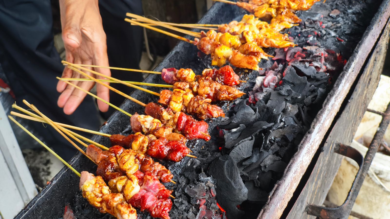 Rows of satay skewers grilling over a long charcoal trough